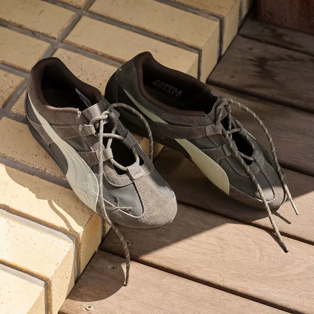 Pair of brown and charcoal PUMA low profile shoes with suede overlays, dark mesh panels, cream Formstrips, and rope laces resting across tiled steps and wooden decking in direct sunlight.