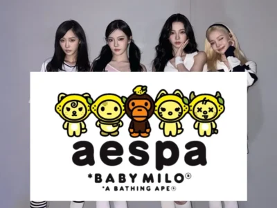 Four members of aespa stand behind a large display board featuring the BABY MILO collaboration artwork, posing against a clean studio backdrop