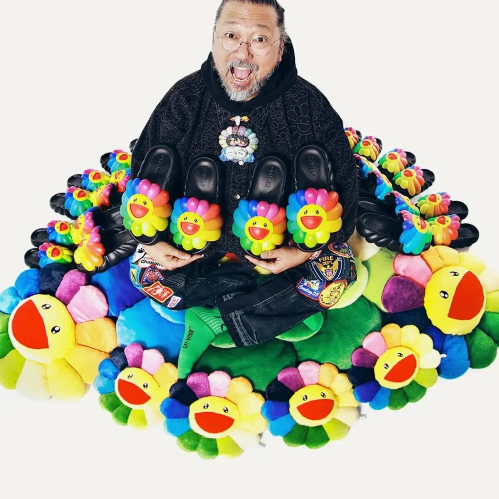 The artist sits cross-legged among dozens of flower cushions and matching slide sandals while holding a pair upright toward the camera. His black outfit with embroidered patches contrasts with the vibrant rainbow accessor