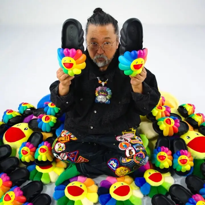 Takashi Murakami sits surrounded by rainbow flower plush toys while holding two black flower-shaped sandals.
