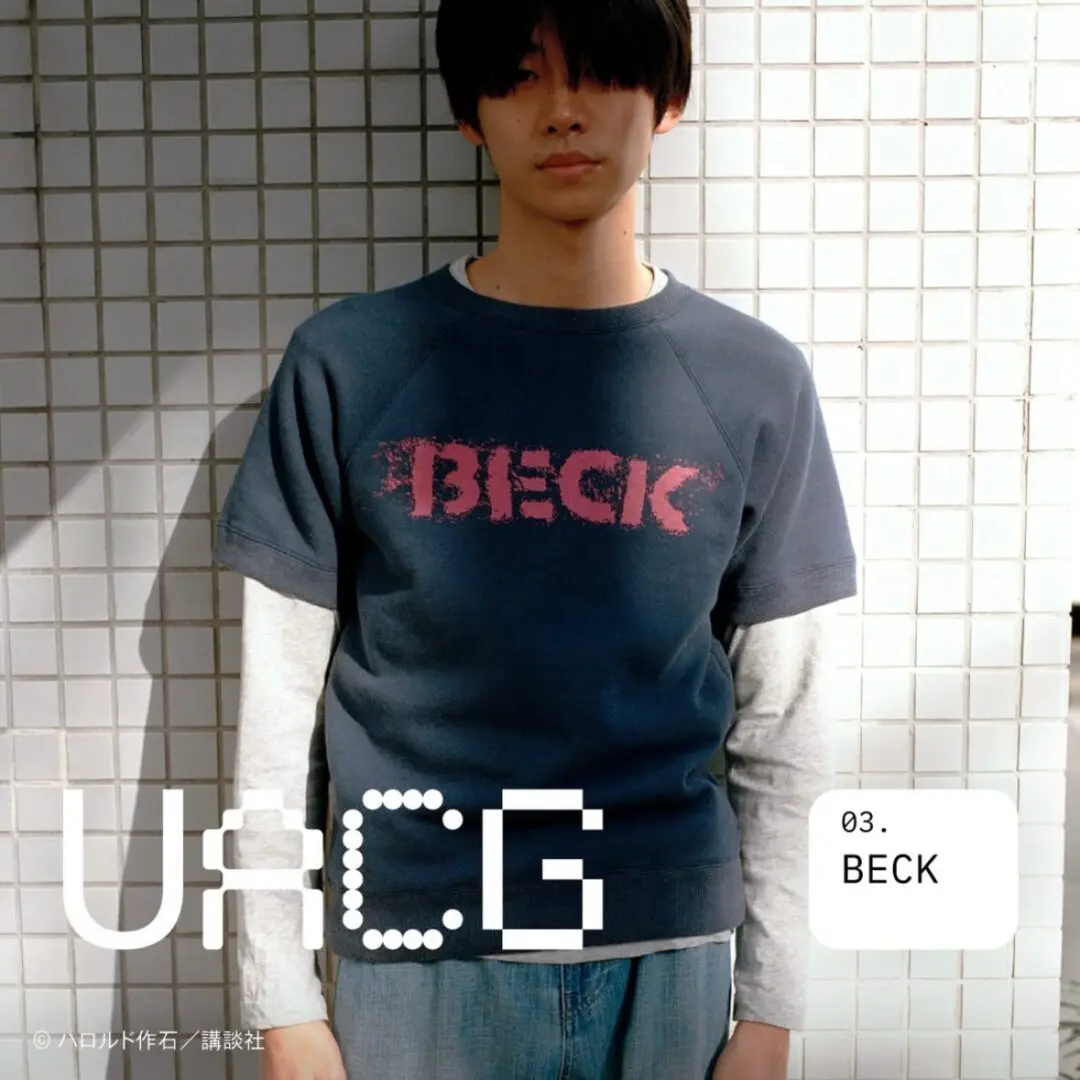 Model wearing a faded blue short-sleeve sweatshirt with a distressed pink BECK logo over a white long-sleeve shirt, paired with light blue jeans against a tiled wall.