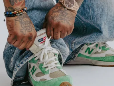 Close-up of tattooed hands tying the laces of mint green and cream New Balance 992 Made in USA sneakers with gum outsoles, paired with faded blue denim, stacked bracelets, and a rose gold wristwatch in a clean studio set