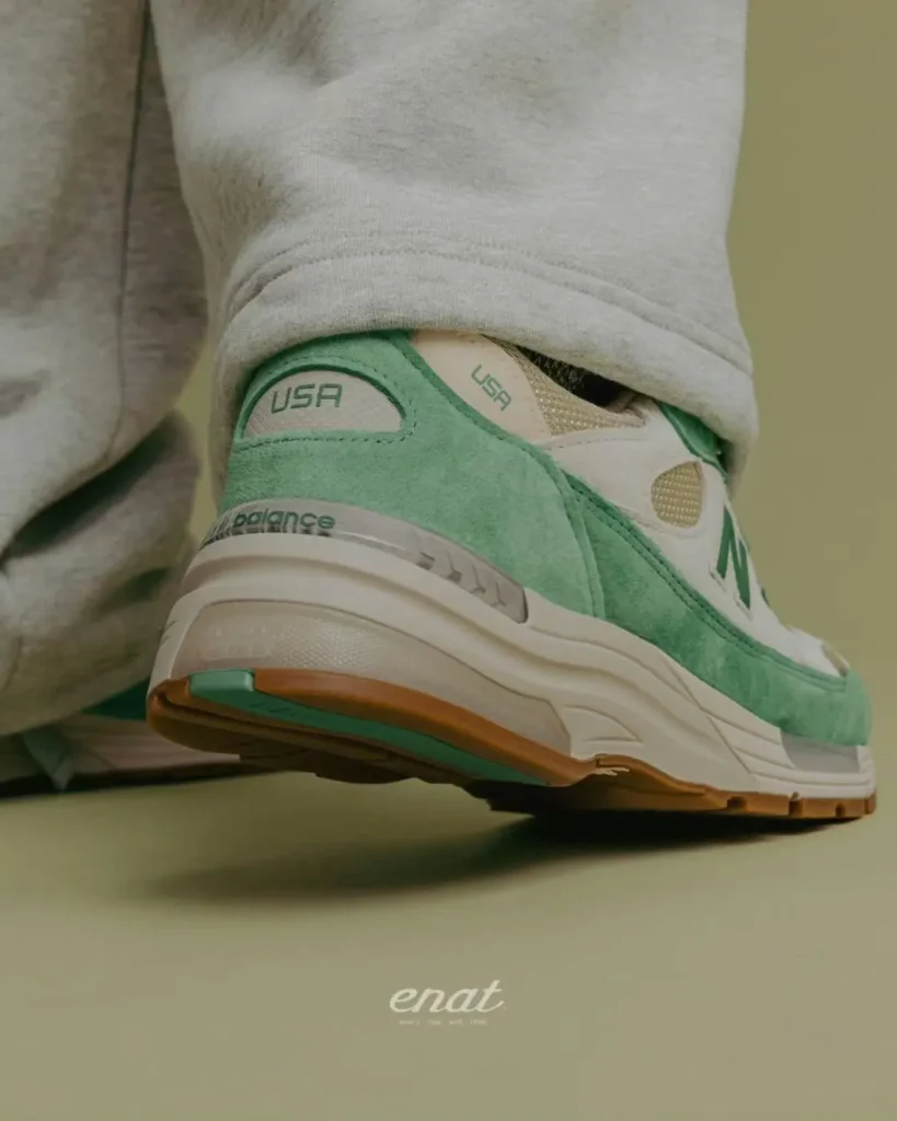Rear three-quarter view of the mint green New Balance 992 Made in USA shoe showing the suede heel panel, embroidered "USA" branding, cream midsole with cushion window, and gum outsole beneath relaxed gray sweatpant