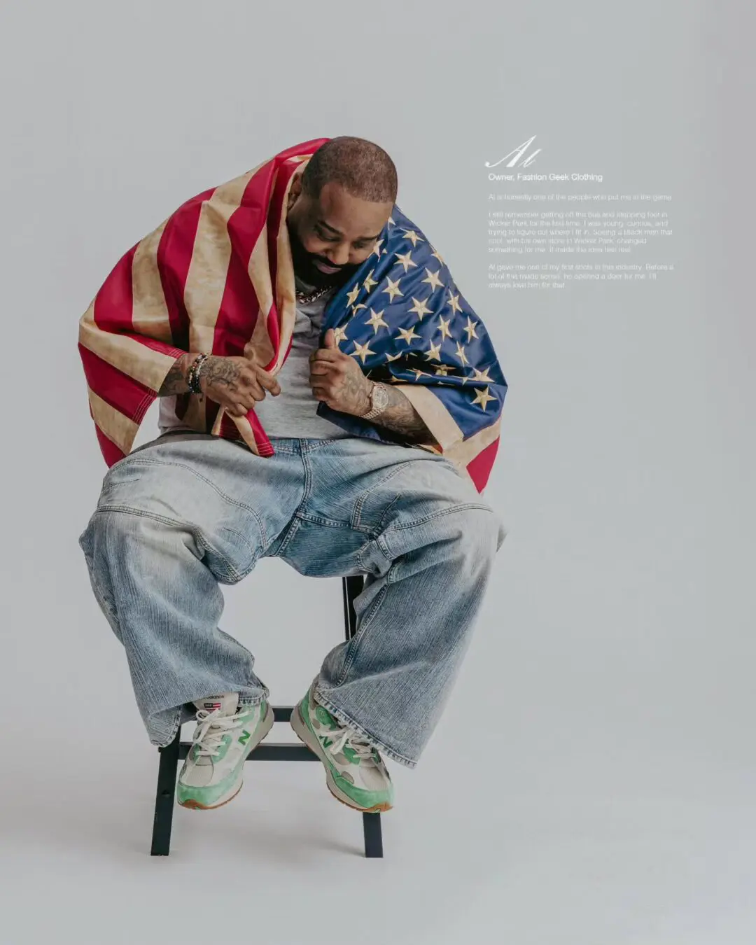 Seated Chicago fashion figure draped in a vintage American flag, wearing oversized light-wash denim and mint green New Balance 992 Made in USA sneakers against a minimalist studio backdrop.