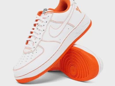 White and orange Nike Air Force 1 Low featuring Kobe Bryant-inspired detailing, orange contrast stitching, white leather upper, bright orange outsole, and stacked studio presentation against a light gray background