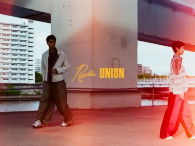 Campaign image of two models wearing Needles x UNION apparel beneath an elevated highway, with the collaboration logo centered against an urban waterfront backdrop