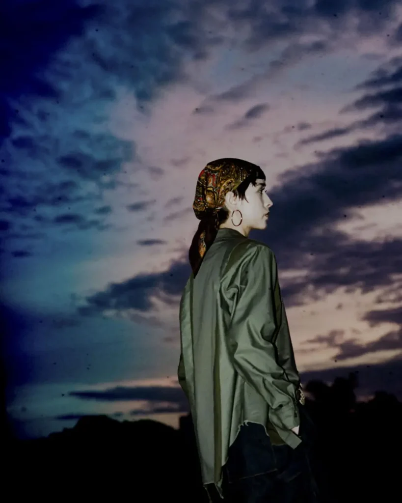 Side-profile portrait of a model wearing a patterned headscarf, olive overshirt, and dark skirt beneath a dramatic twilight sky