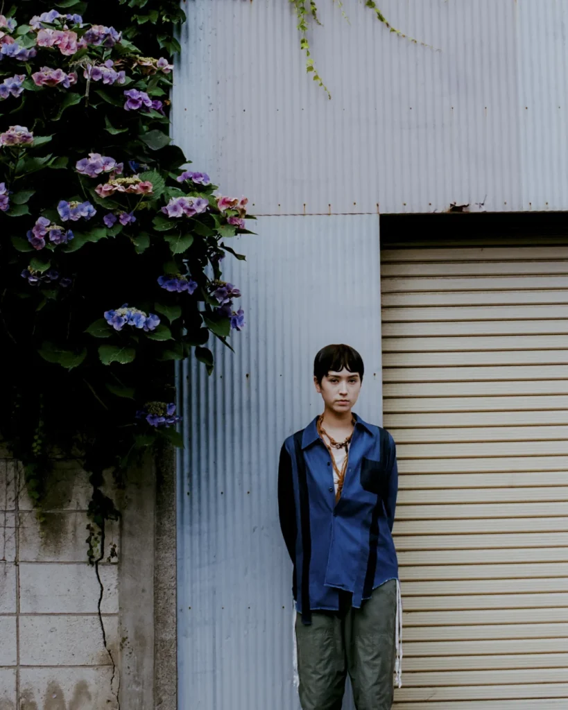 Model standing in front of a corrugated metal wall and blooming hydrangeas wearing a blue Needles x UNION overshirt, layered necklaces, and olive cargo trouser
