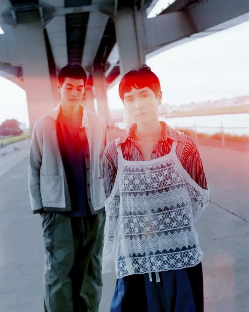 Two models wearing Needles x UNION apparel pose beneath an overpass, combining lace layering, textured jackets, oversized trousers, and soft red light leaks for a cinematic editorial look
