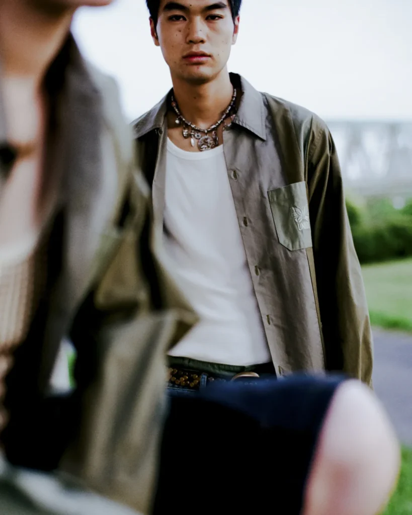 Close-up portrait of a model wearing an olive Needles x UNION overshirt layered over a white tank top, finished with layered necklaces and relaxed denim