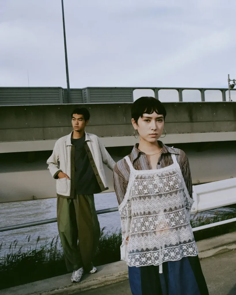 Two models pose beside a riverside barrier wearing relaxed Needles x UNION outfits, featuring lace layering, wide-leg trousers, textured outerwear, and patterned shirt