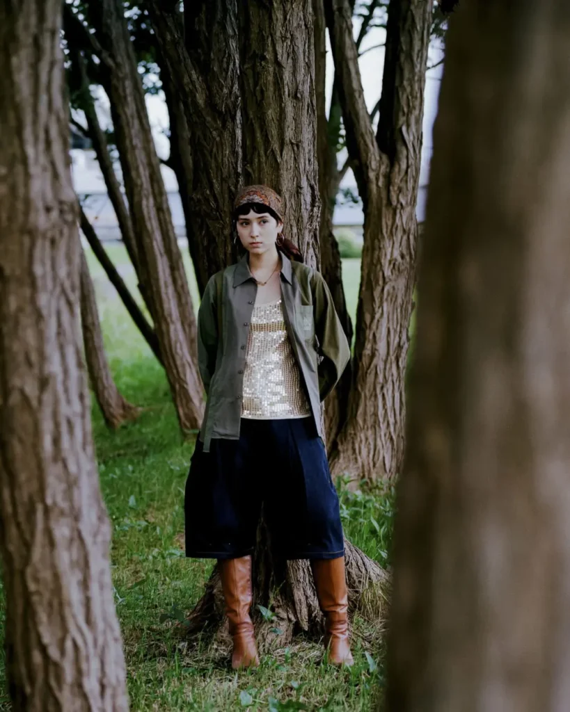 Model standing among tall trees wearing a green Needles x UNION overshirt, sequined top, dark culottes, brown knee-high boots, and a patterned headscarf