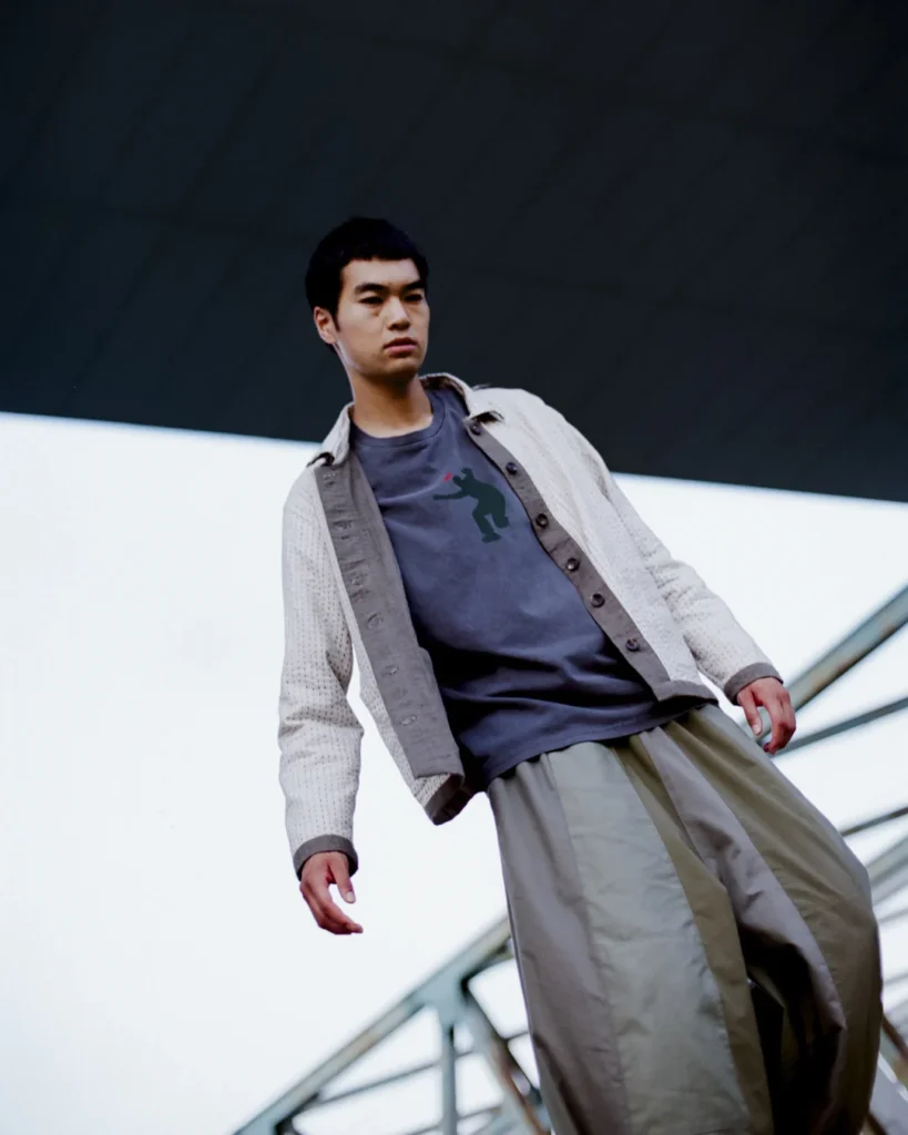 Low-angle portrait of a model wear a textured cream jacket layered over a graphic Tee with oversized pleated trousers in a modern architecture set