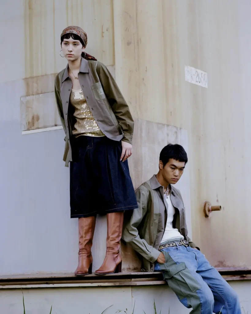 Two models styled in coordinated Needles x UNION pieces pose against an industrial building, pairing utility overshirts, relaxed denim, sequined layers, and knee-high boots