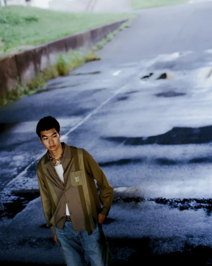 Model wearing an olive patchwork Needles x UNION overshirt, white tank top, layered necklaces, and cargo-style denim posed against a projected urban street scene