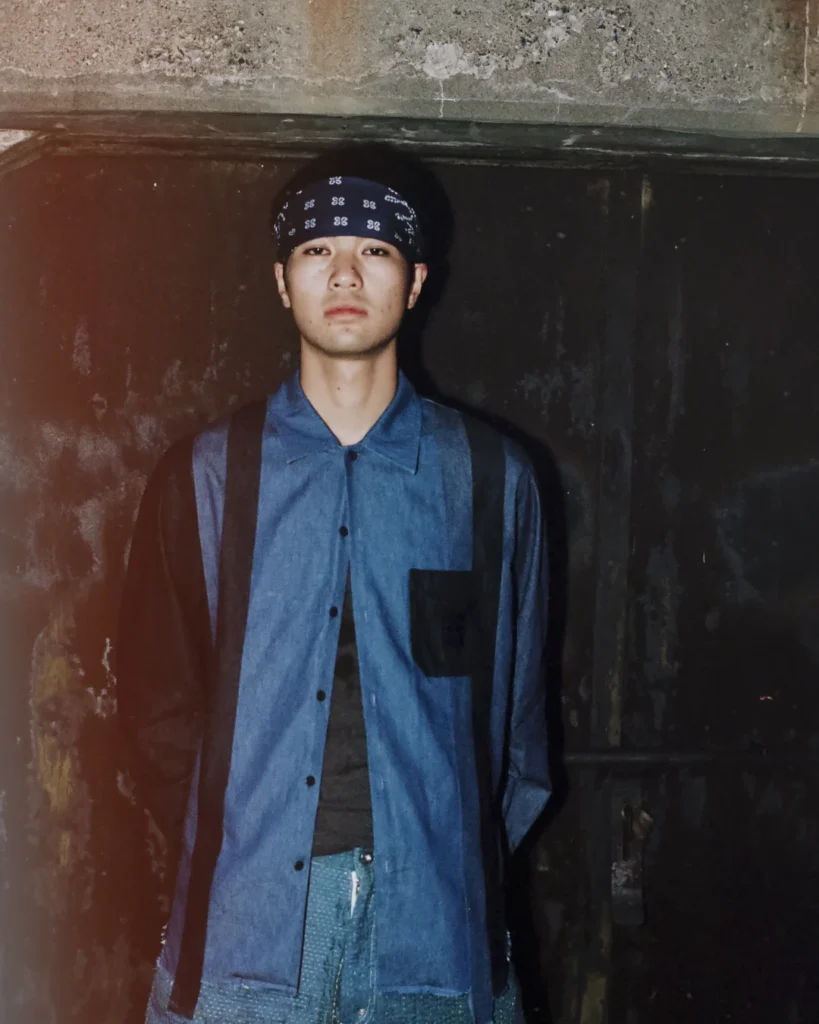 Portrait of a model wearing a blue Needles x UNION overshirt with black panel detailing, a patterned bandana, and textured trousers against a dark industrial backdrop