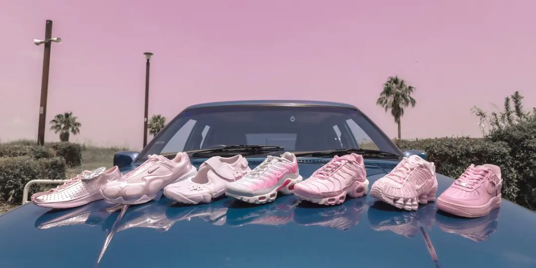 Six pastel pink Nike sneakers arranged across the hood of a vintage blue car beneath a soft pink sky, showcasing a mix of classic and performance silhouettes including Air Max, Shox, Air Force 1, and retro running designs.