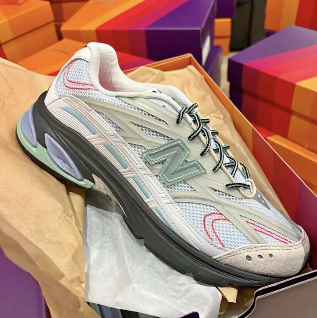 A New Balance running shoe in white mesh with translucent overlays, a mint green "N" logo, black rope laces, pastel blue accents, red embroidered details, and a sculpted black midsole, displayed inside an open shoebox with colorful New Balance boxes stacked in the background