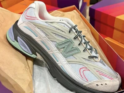 A New Balance running shoe in white mesh with translucent overlays, a mint green "N" logo, black rope laces, pastel blue accents, red embroidered details, and a sculpted black midsole, displayed inside an open shoebox with colorful New Balance boxes stacked in the background