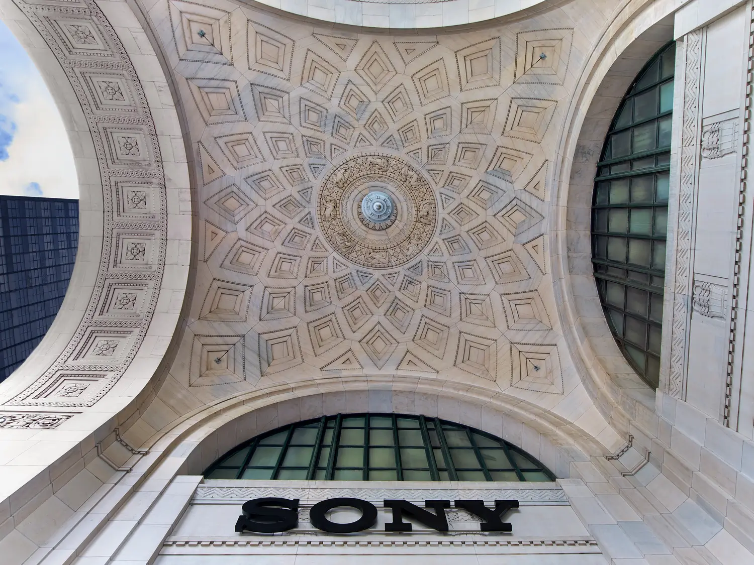 Looking upward beneath a grand stone archway with an ornate coffered ceiling, decorative geometric carvings, and a large SONY sign mounted above the entrance.