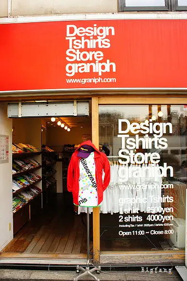 Graniph retail storefront with a bright red 'Design Tshirts Store graniph' sign, large display windows showcasing graphic apparel, and a mannequin dressed in a colorful Graniph T-shirt outside the entrance.