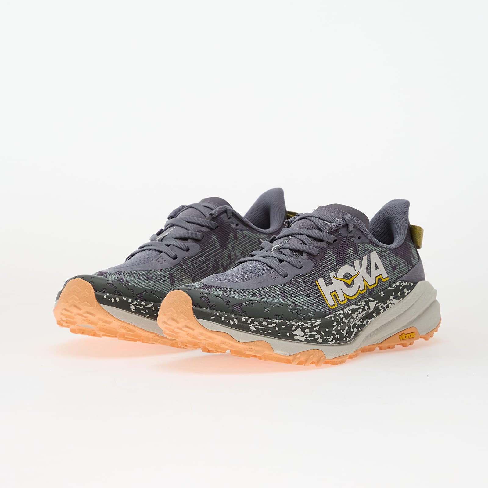 Angled front three-quarter view of a pair of HOKA trail running shoes in dark gray with oversized white and yellow HOKA branding, textured striped mesh uppers, speckled mudguards, thick cream midsoles, and peach-colored Vibram rubber outsoles on a white background.