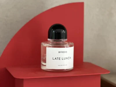 Clear glass bottle of BYREDO Late Lunch Eau de Parfum displayed on a sculptural matte red pedestal with a curved back panel. The fragrance features BYREDO's signature black dome cap and minimalist white label, photographed against a warm neutral plaster wall in soft natural light
