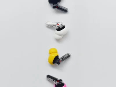Black, white, yellow, and pink Nothing Ear (3a) wireless earbuds are arranged vertically on a light gray background, highlighting the transparent stems and bold color option