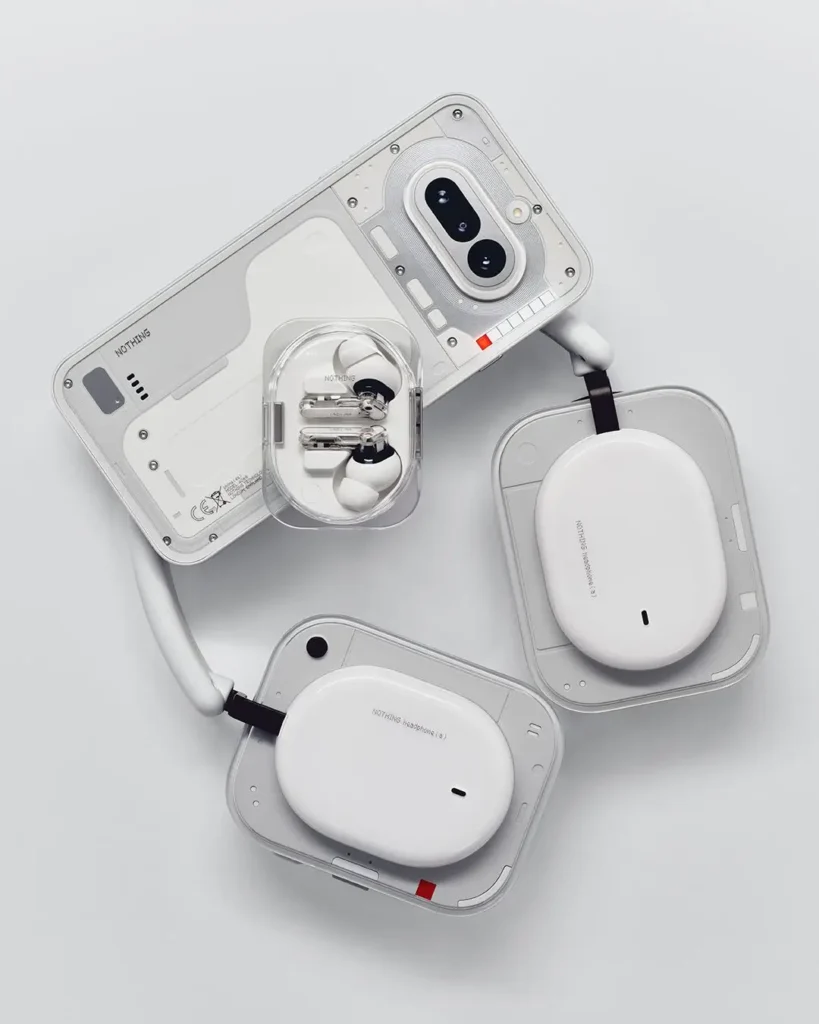 A white Nothing smartphone, matching over-ear headphones, and white Nothing Ear (3a) earbuds in a transparent charging case are arranged together on a light gray background to showcase the brand's ecosystem