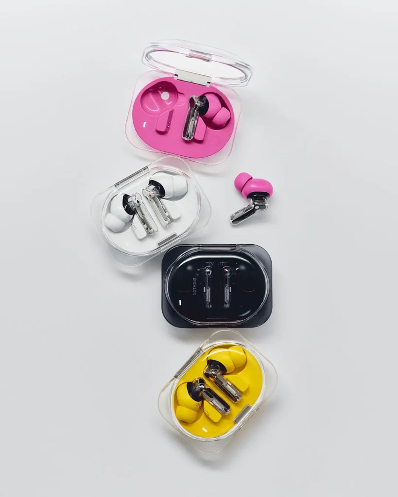 Black, white, yellow, and pink Nothing Ear (3a) charging cases are stacked vertically on a neutral background, with one pink earbud placed outside its case to highlight the available color option