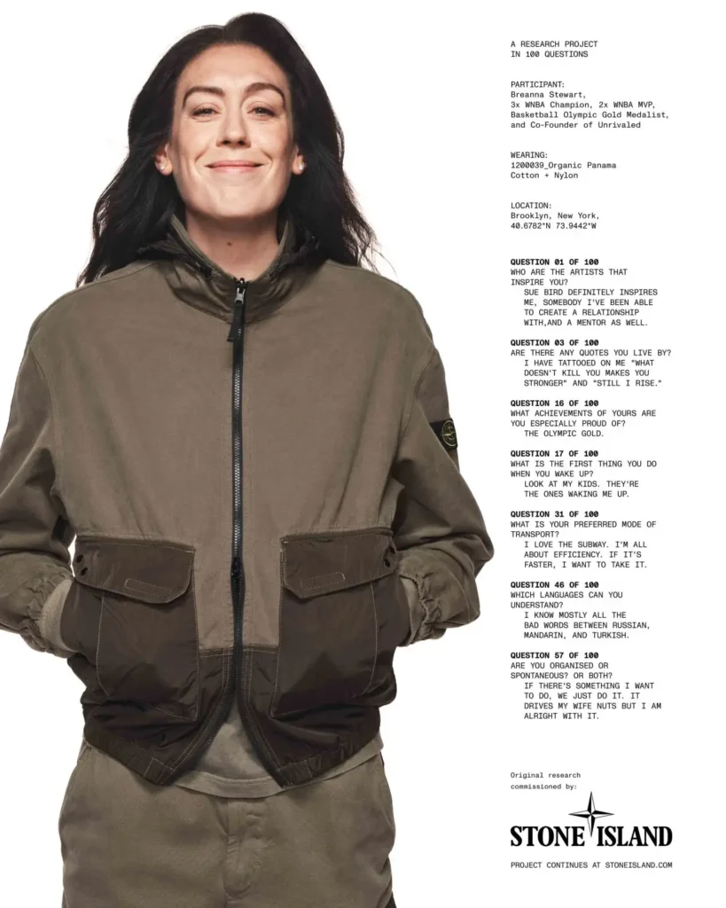 Breanna Stewart smiles while posing against a white studio backdrop wearing a two-tone Stone Island zip-front jacket with the brand's signature compass badge on the left sleeve, hands tucked into the front pockets. A column of printed interview questions and answers appears beside her, outlining her achievements, inspirations, personal philosophy, and details about the featured Organic Panama Cotton + Nylon garment as part of Stone Island's "A Research Project in 100 Questions" campaign