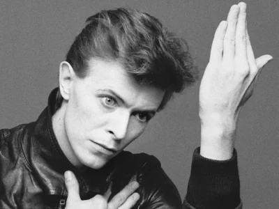 A black-and-white studio portrait of David Bowie with a sharp side-shaved hairstyle wearing a black leather jacket. They hold one hand against their chest while raising the other vertically beside their face, looking directly into the camera with an intense expression. The minimalist composition and high-contrast lighting evoke a classic late-1970s portrait style