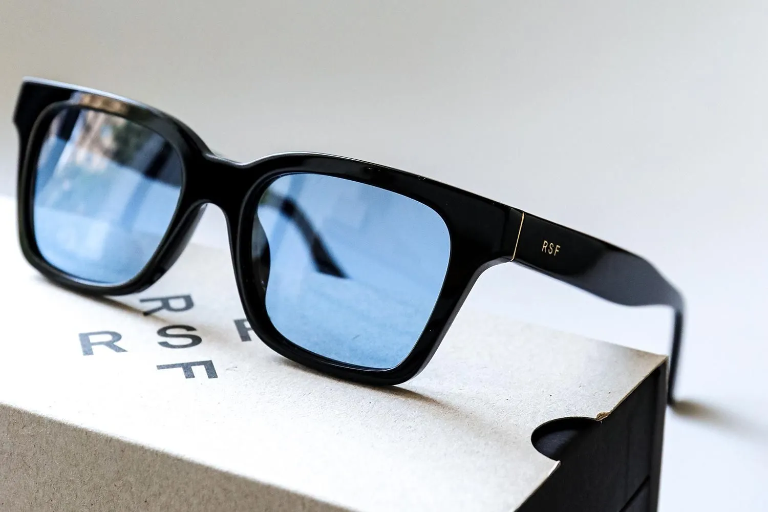 RSF Blades Series 2026 sunglasses with a glossy black acetate frame and blue-tinted lenses resting on an RSF-branded box, highlighting the minimalist temple logo and handcrafted finish.