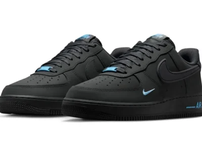 Pair of black Nike Air Force 1 Low shoe shown in a three quarter front angle on a white background, featuring matte black leather uppers, tonal oversized Swooshes, and bright blue accents on the lace dubrae, mini embroidered Swoosh, tongue branding, and midsole AIR logo