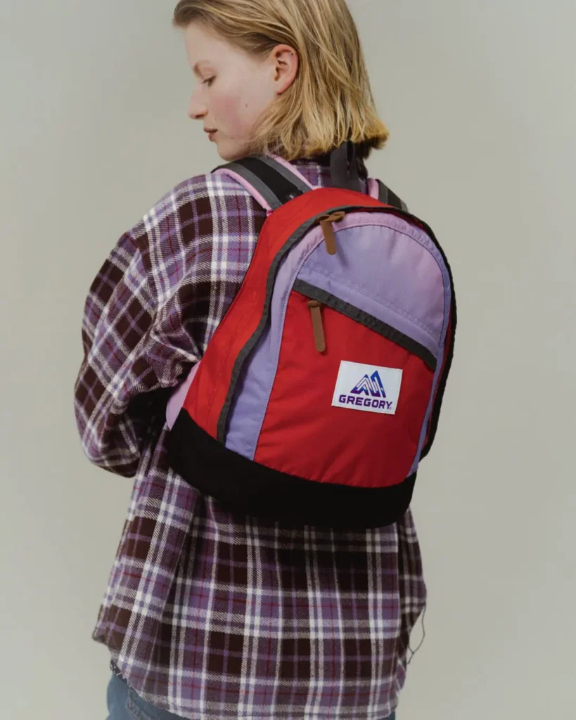 Model wearing the Gregory × BEAMS BOY backpack, featuring a red body with lavender panels, black base, leather zipper pulls, and adjustable shoulder straps paired with a purple plaid overshirt for a heritage-inspired outdoor look