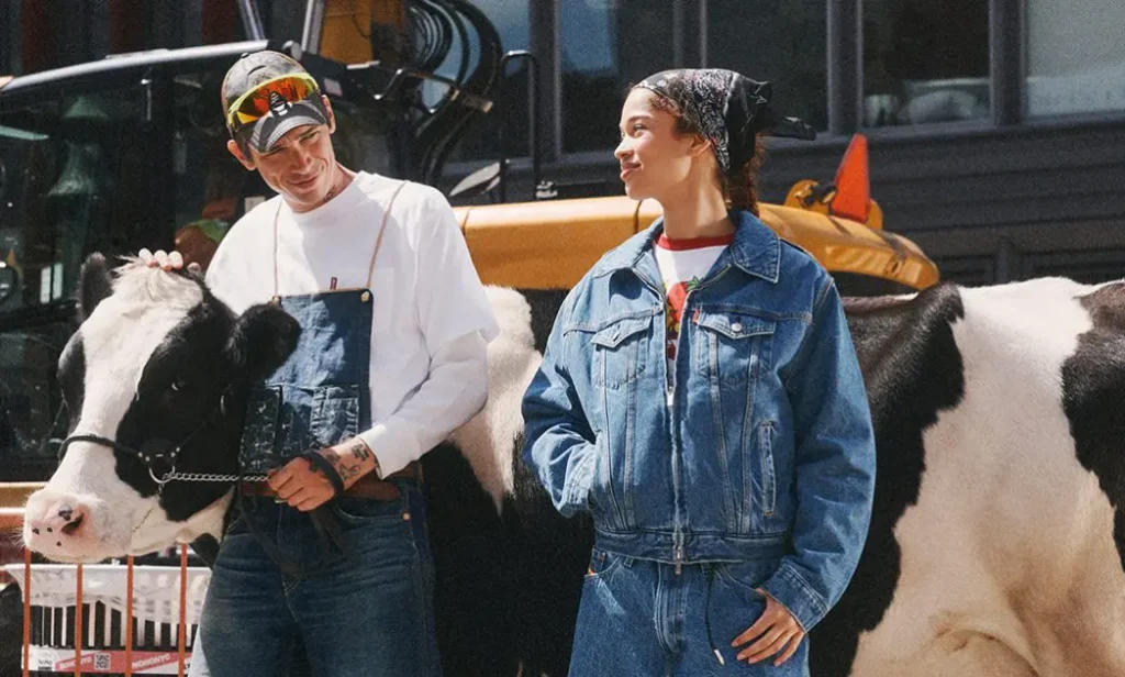 Two models in Levi's x Sky High Farm denim stand beside a black-and-white dairy cow on a city street, blending workwear-inspired fashion with an unexpected urban farm set