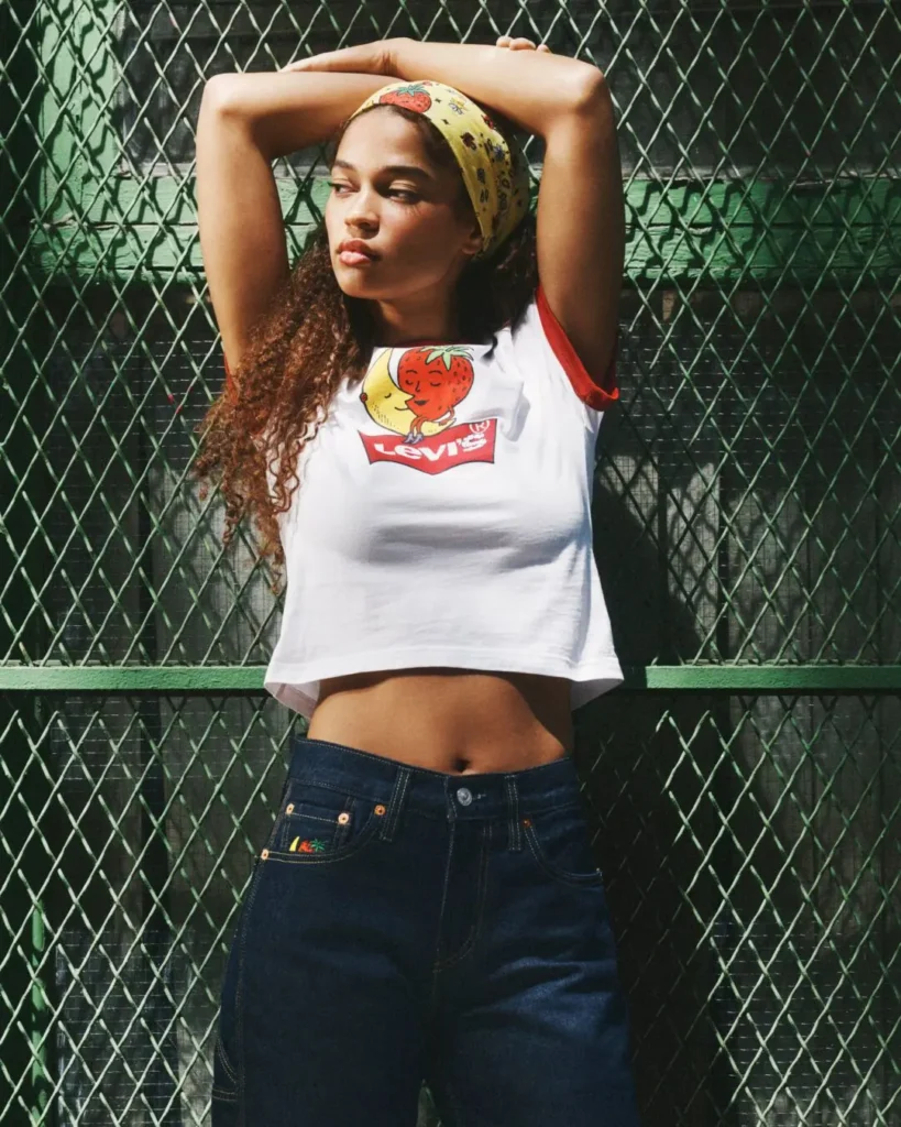 Model posing against a green chain-link fence wearing a white ringer T-shirt with a Levi's logo and Sky High Farm strawberry-and-moon graphic, paired with dark indigo denim and a matching printed bandana