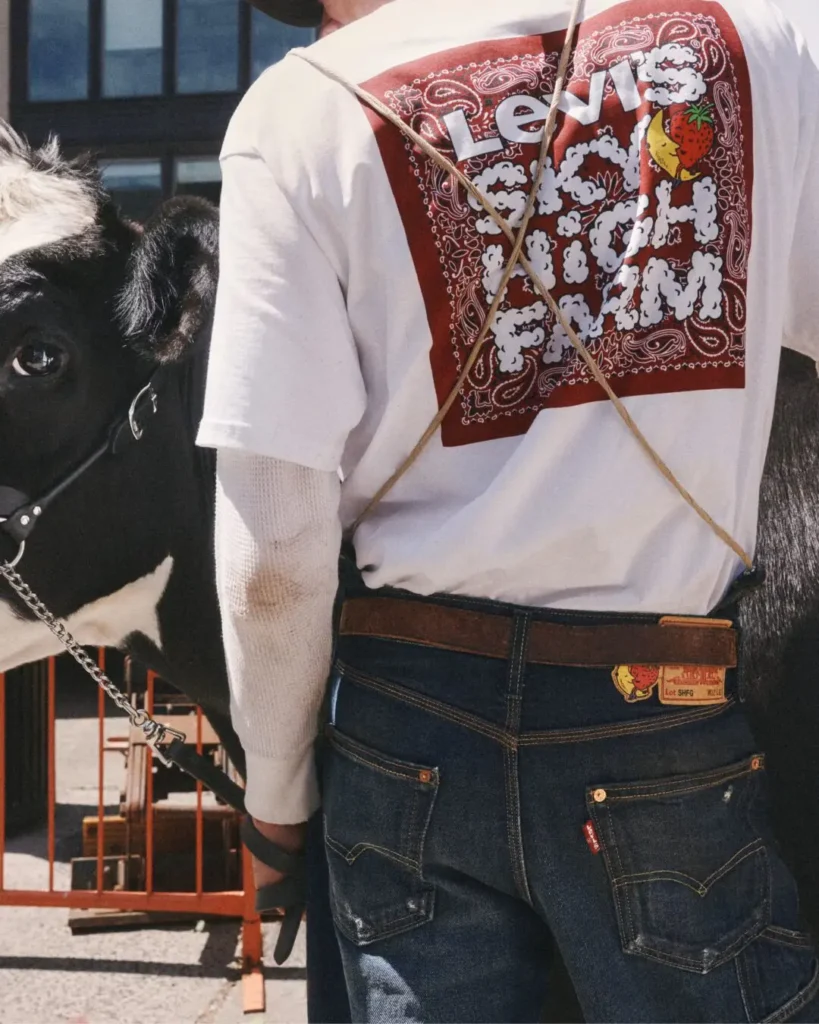 Rear view of a model in dark indigo carpenter jeans and a white graphic Tee with a large red bandana-inspired Sky High Farm print, standing beside a dairy cow