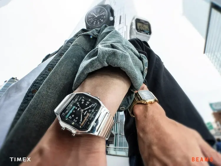Campaign image of the Timex × BEAMS collaboration showing the new analog-digital watch in polished stainless steel on a wrist, photographed alongside classic Timex models in an urban setting.