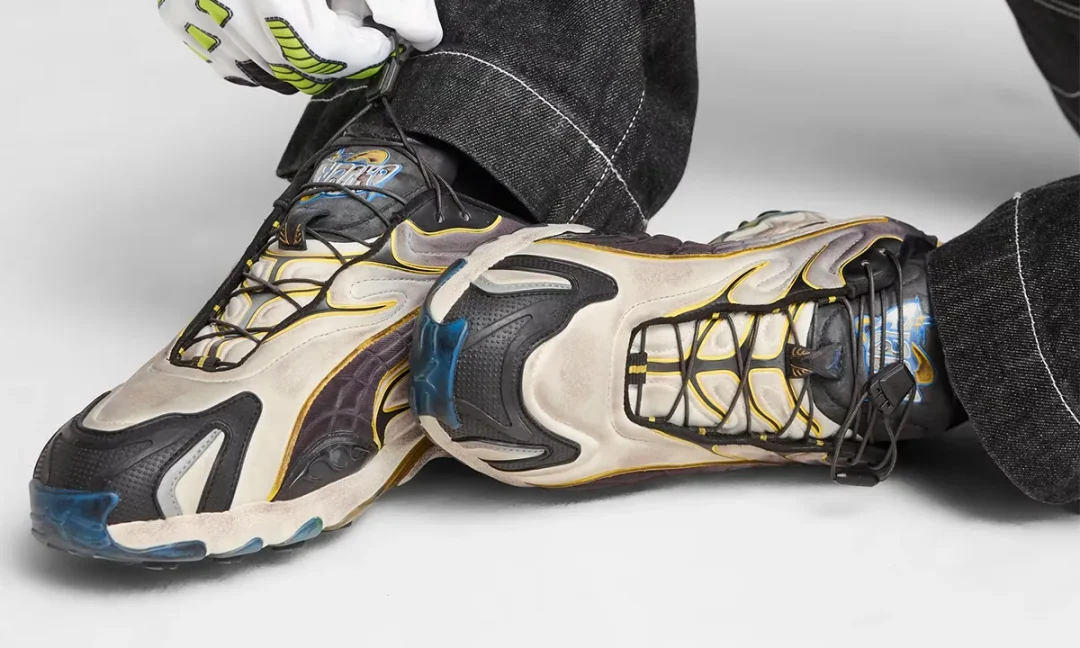 A close up of the collaborative PUMA Inhale sneakers worn with wide leg black denim and fluorescent riding gloves. The shoes feature a weathered silver upper, black overlays, yellow piping, blue translucent outsole accents, and a quick lace system that reinforces their worn in motorsport aesthetic.