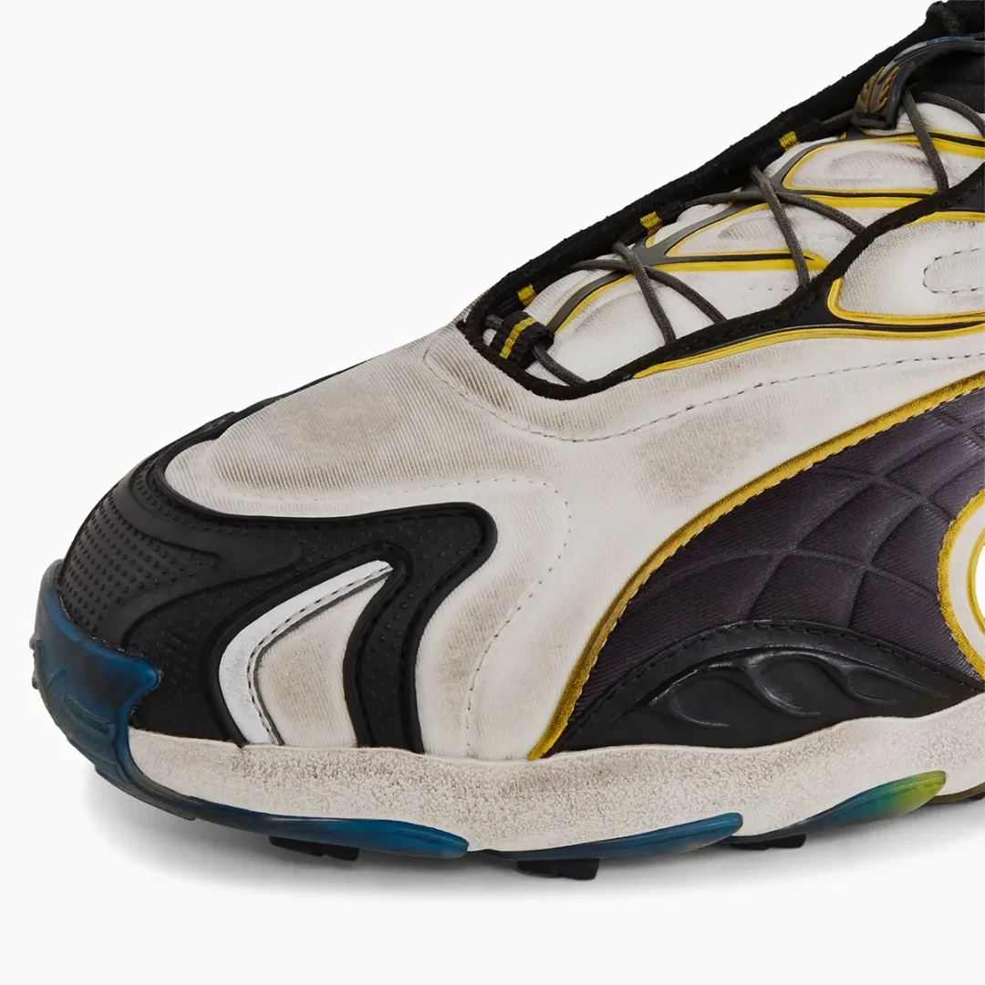 A macro view of the PUMA Inhale sneaker highlights its distressed silver textile toe box, textured black rubber mudguard, quilted dark side panel, yellow piping, weathered sculpted midsole, and translucent blue outsole accents that reinforce the intentionally worn racing inspired finish.