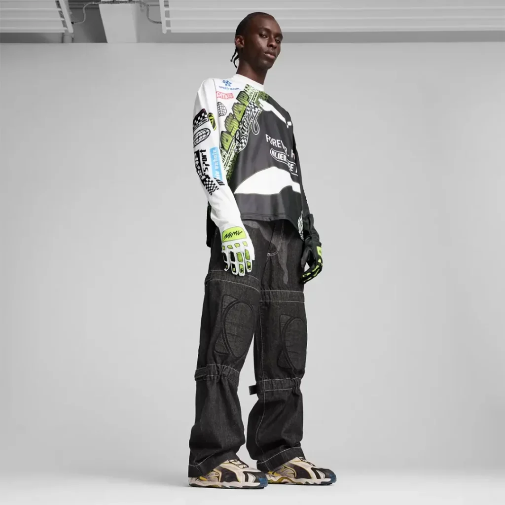 A model stands in a studio wearing the collaborative PUMA sneakers with oversized black denim pants, a graphic motocross inspired jersey, and fluorescent riding gloves. The look emphasizes motorsport influences and technical streetwear style