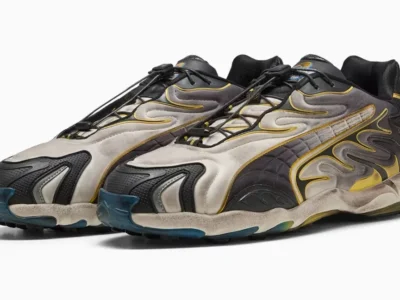 A three quarter product view of the PUMA sneakers highlights the sculpted silhouette, weathered silver textile upper, quilted side panels, flowing black overlays, yellow trim, and translucent blue outsole accents against a white background