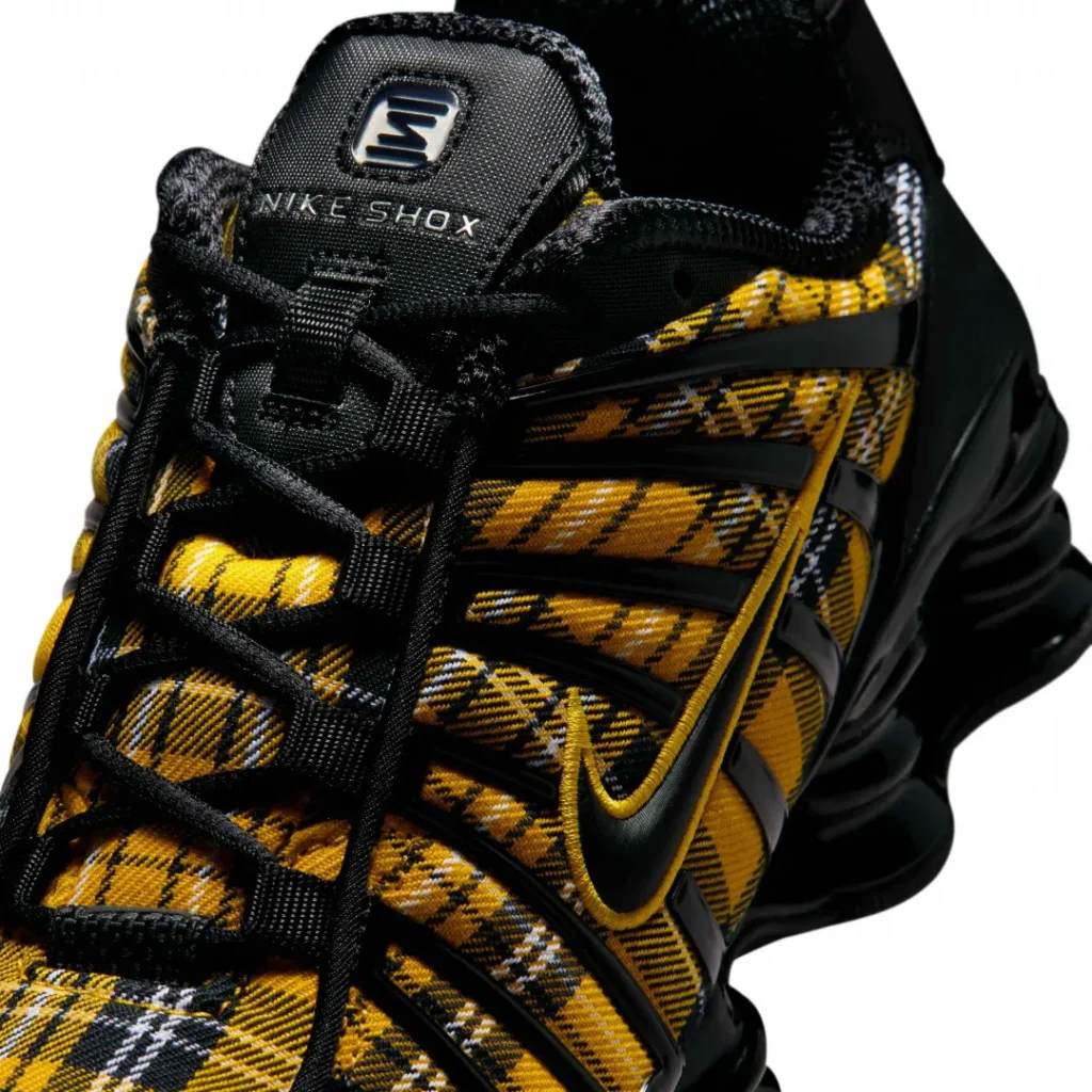 Close-up of the Nike Shox shoe highlighting the yellow tartan plaid upper, black TPU cage, embroidered yellow Swoosh, black laces, and Nike Shox tongue brand