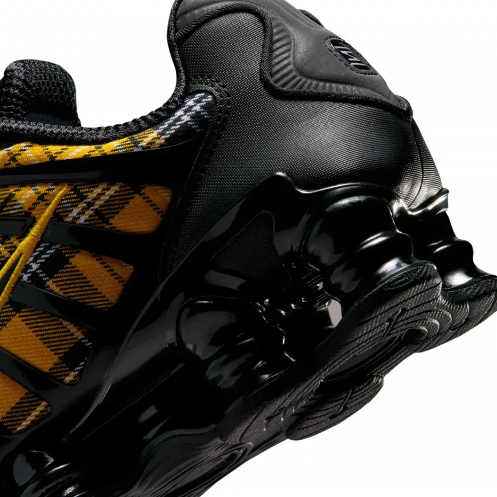 Close-up of the Nike Shox heel showcasing the glossy black Shox cushioning columns, black heel panel, and tartan plaid textile wrapping the rear quarter