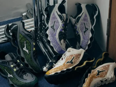 A group of Mizuno Wave Prophecy LS sneakers in green, purple, and gold-accented colorways are arranged on a blue carpet beside polished chrome furniture, highlighting the sculpted Wave sole and layered mesh upper