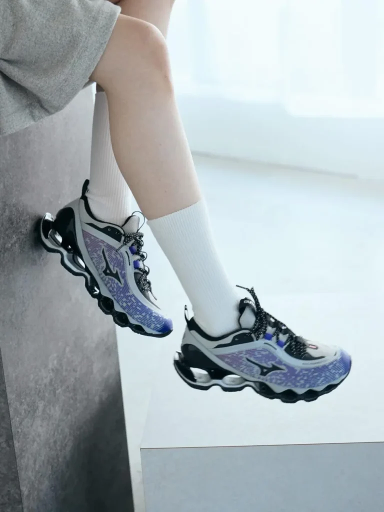 A person wearing the purple Mizuno Wave Prophecy LS sits on a ledge with white crew socks, showcasing the shoe's gradient mesh upper, black Wave midsole, and reflective details in natural light
