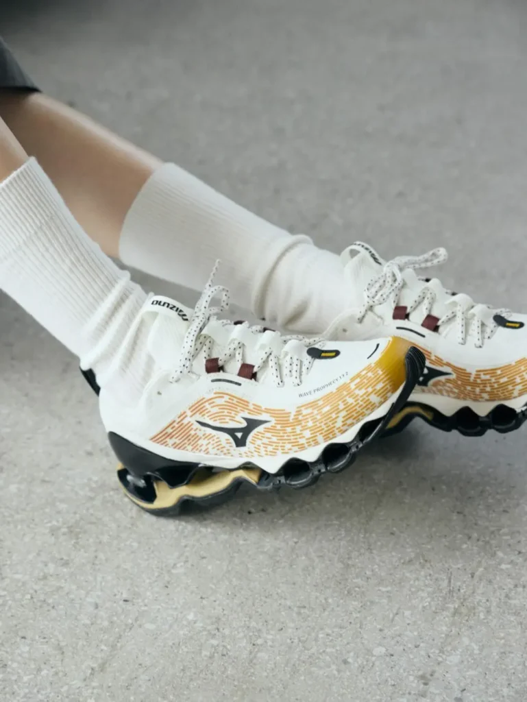 A close-up of the white and gold Mizuno Wave Prophecy LS on foot, featuring a white engineered mesh upper with amber gradient graphics, speckled laces, and the signature hollow Wave cushion system