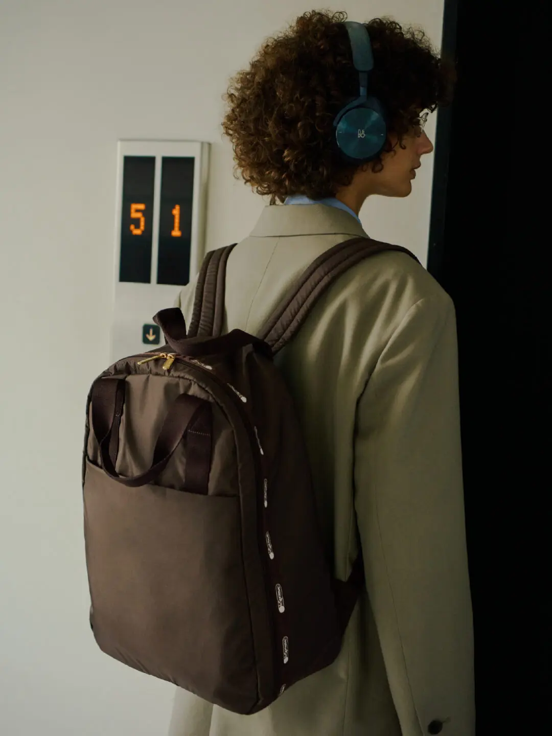 Person wearing a brown LeSportsac backpack with dual top handles and padded shoulder straps while waiting by an elevator, highlighting the bag's clean, lightweight commuter design.