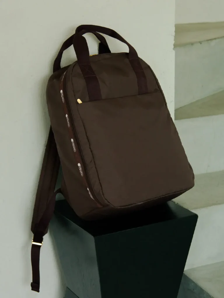 Brown LeSportsac backpack leaning against a modern pedestal, emphasizing its streamlined profile, front pocket, and min
